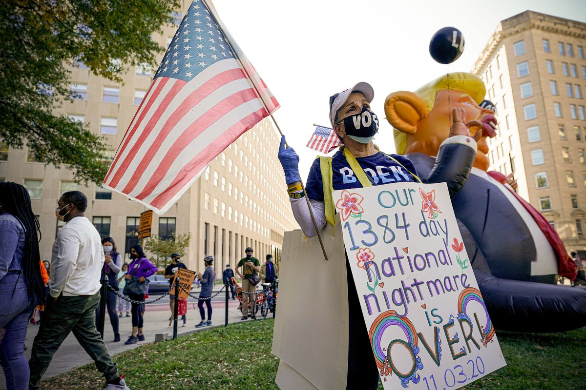 A protester in a vote mask and Biden t-shirt holds a US flag and wears a sandwich board that reads “Our 1384 day national nightmare is over.” The board is decorated with flowers and rainbows.