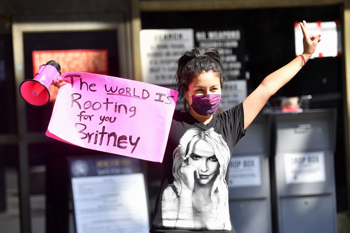 A masked woman in a Britney Spears T-shirt carries a pink bullhorn and holds a sign saying, “The world is rooting for you, Britney.”