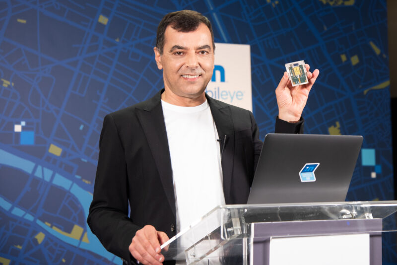 A man at a podium smiles while holding up a palm-sized computer component.