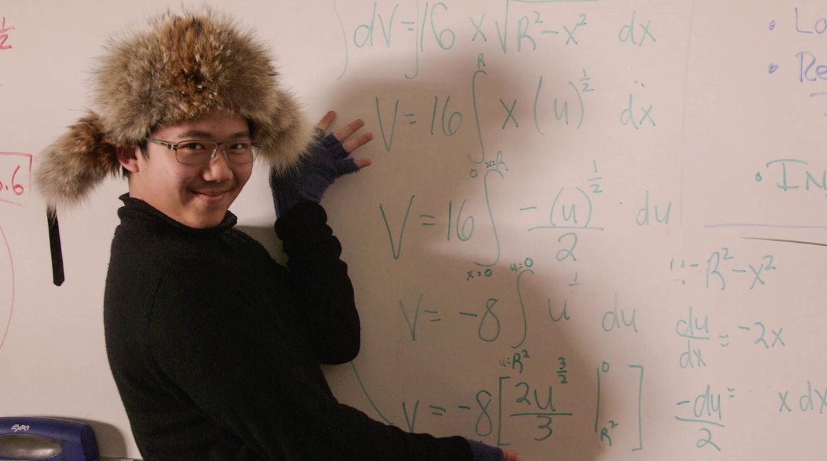 A teenager in a fuzzy cap stands in front of a whiteboard gesturing with a smile at calculus equations.