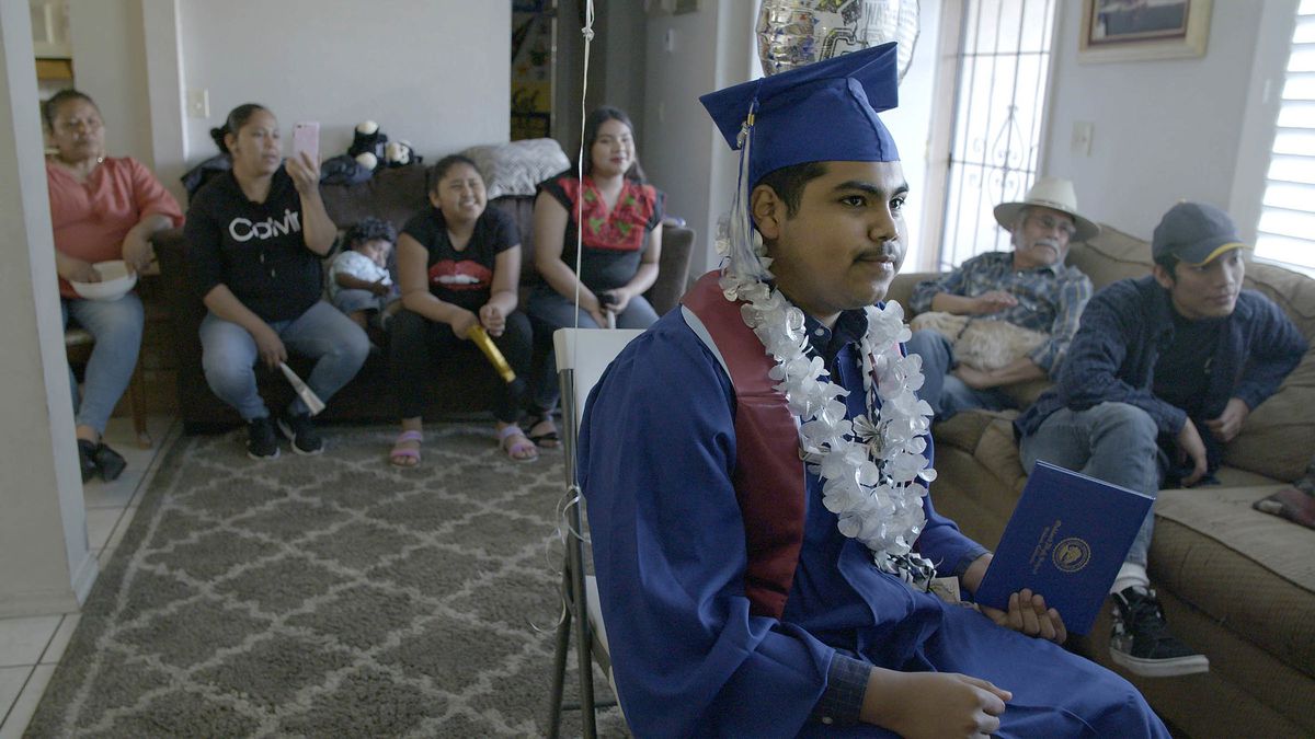 A teenager sits in his graduation gown in his living room, surrounded by family members.