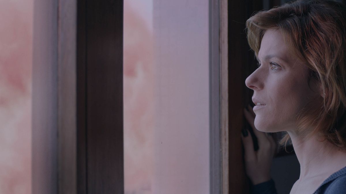 A woman stands at a window, looking at a pink cloud outside.