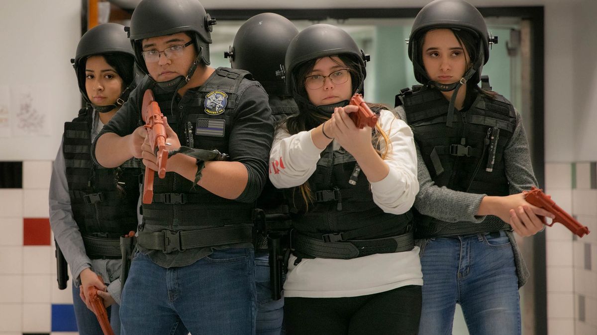 A group of high school students in riot police garb clusters in a hallway, holding fake plastic guns.