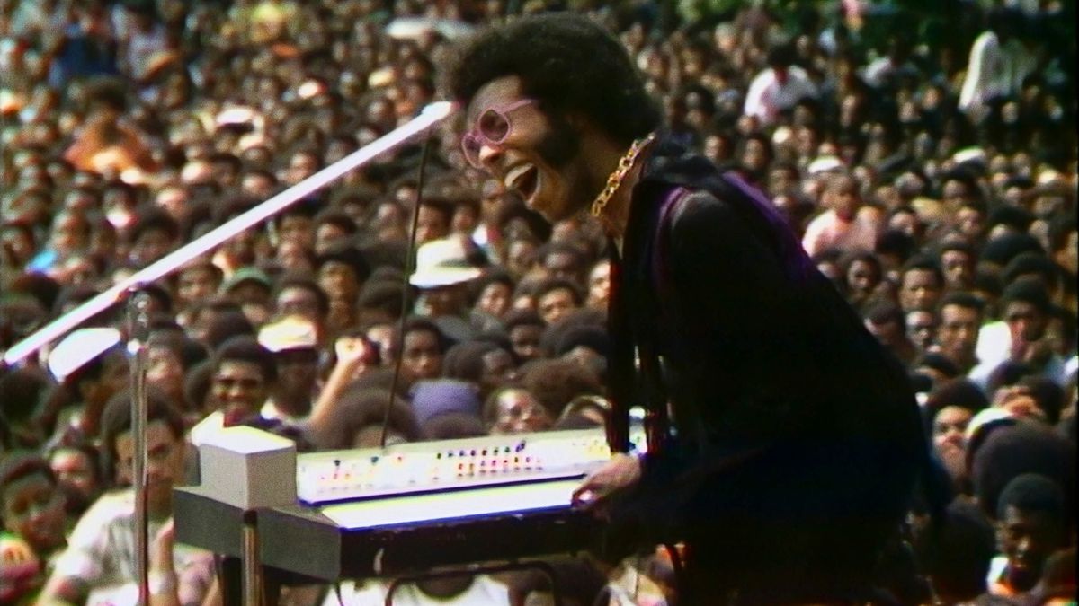 Sly Stone sings into a microphone in front of a keyboard and a crowd.